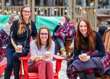 Three women smiling and holding coffee cups while sitting on red Adirondack chairs outdoors, with cozy blankets and string lights in the background.