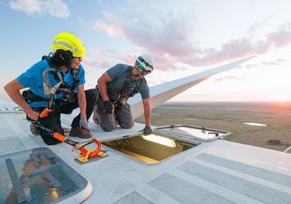 Two workers wearing safety gear and helmets on top of a wind turbine at sunset, inspecting an open hatch.