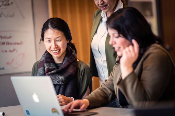 Three women in an office setting collaborate around a laptop, smiling and engaged in discussion.