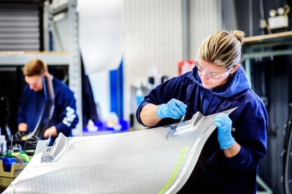 A worker in safety goggles and blue gloves carefully inspects a metallic component in a manufacturing facility, with another worker focused in the background.