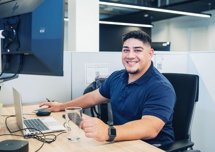 A smiling office worker in a navy polo shirt sits at a desk with a laptop and monitor, holding a glass and working in a modern, bright workspace.