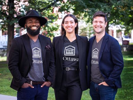 EThree smiling professionals in matching blazers with 'Ottawa' logos stand outdoors in a park-like setting.