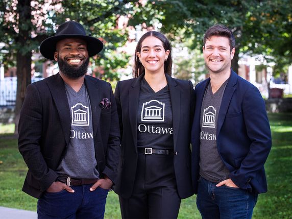 Three smiling professionals in matching blazers with 'Ottawa' logos stand outdoors in a park-like setting.