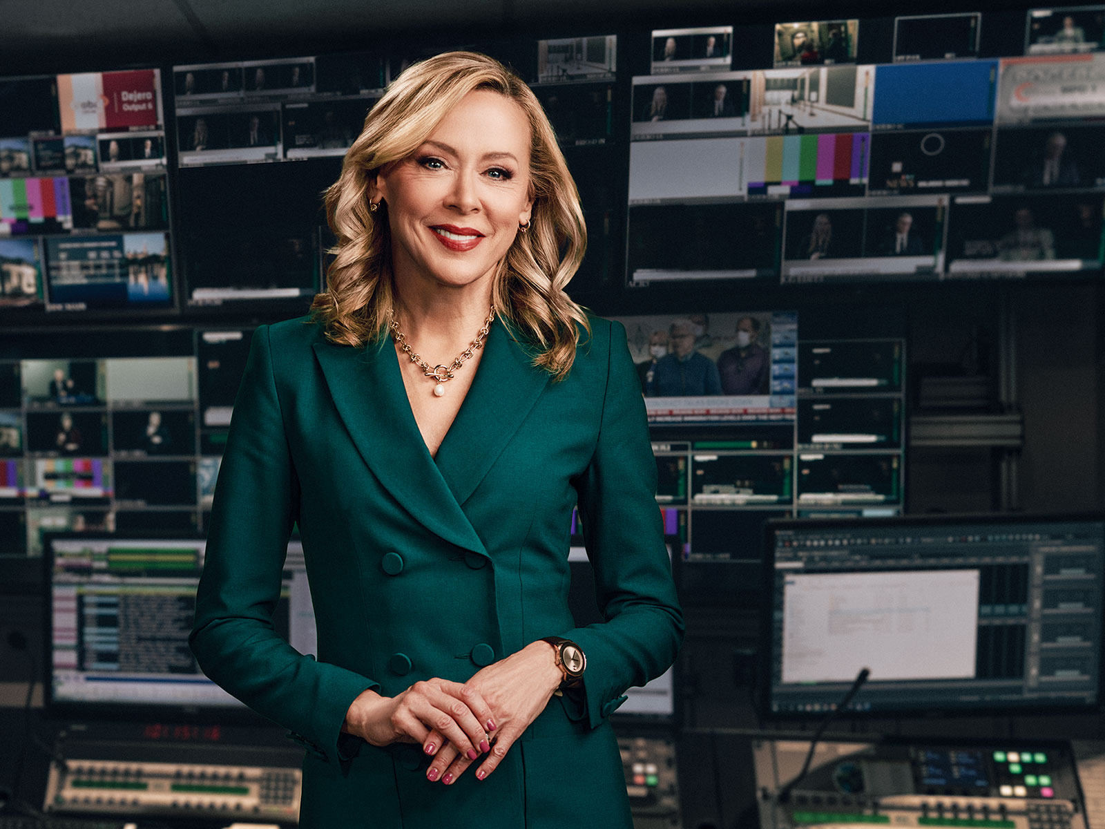 A confident woman in a green blazer with a beaming smile stands in a broadcast control room filled with screens and technical equipment.