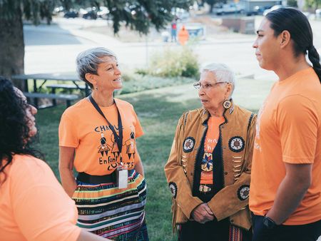 Four people are engaging in conversation outdoors, two women and one man wearing orange shirts with a slogan, 'Every Child Matters,'' and an elder in a decorated traditional jacket, with trees and a park setting in the background.