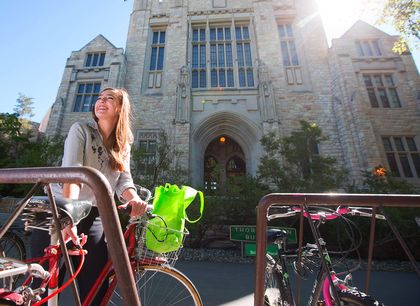 A smiling woman with a bicycle near a historic stone building with large windows and an arched entrance.