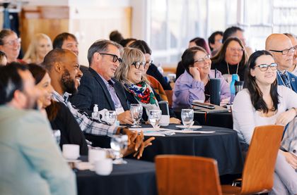 A diverse group of people seated at round tables, smiling and engaged during a conference or presentation.