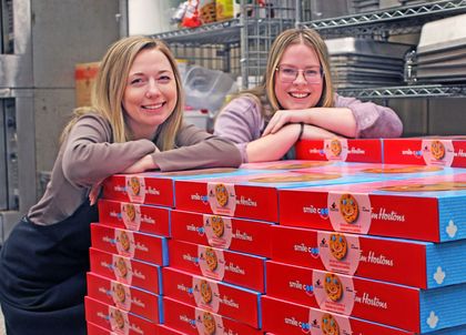 Two smiling women lean on a stack of Tim Hortons Smile Cookie boxes in a kitchen or storage area.