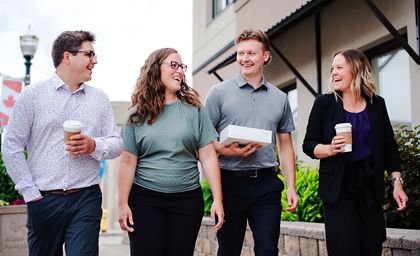 Four professionally dressed people walking outside, smiling and holding coffee cups and a box.