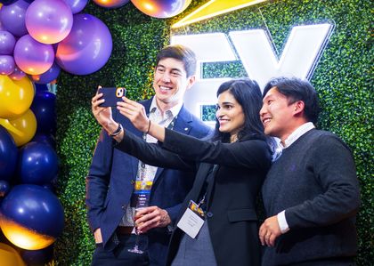 Three professionals pose for a selfie in front of a vibrant backdrop featuring balloons and illuminated signage, capturing a moment of camaraderie at an EY event.