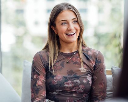 A smiling young woman wearing a floral-patterned top and a necklace with a 'D' pendant, sitting in a bright, airy space with a blurred green backdrop.